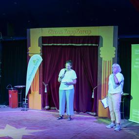 Zwei Frauen stehen auf einer Zirkusbühne, eine spricht ins Mikrofon, im Hintergrund ein rotes Vorhang und Sponsorenbanner.