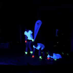 Drei Personen in leuchtenden Shirts und Armbändern in einem dunklen Raum bei einer Aufführung oder Übung.