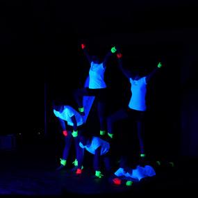 Fünf Personen in weißen T-Shirts und leuchtenden Handgelenkbändern stehen in einem dunklen Raum in verschiedenen Posen.