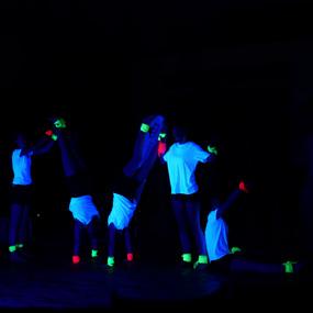 Fünf Personen in weißen Shirts führen im Dunkeln akrobatische Bewegungen mit fluoreszierenden Accessoires aus.