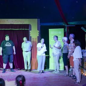 Gruppe von Personen steht auf einer Bühne im Zirkuszelt, während eine Frau spricht und die anderen zuhören.