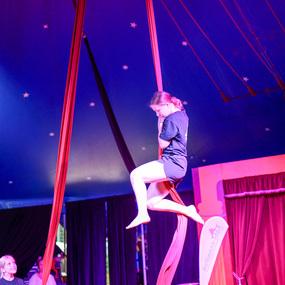 Mädchen auf roten Tuchbändern im Zirkuszelt, während ein anderes Kind zuschaut. Beleuchtung in lila und blauen Tönen.