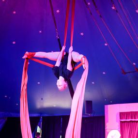 Akrobatin in roten Tüchern schwingt kopfüber in einem Zirkuszelt mit lila Himmel und Sternen.