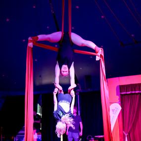 Zwei Akrobatinnen in einem Zelt, eine hängt kopfüber an roten Tüchern, die andere greift sie von unten.