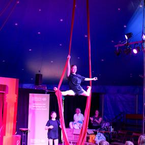 Ein Mädchen führt eine Akrobatik-Nummer an roten Tüchern unter einem bunten Zirkuszelt auf, umgeben von Zuschauern.