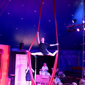 Akrobatin schwebt an roten Tüchern und vollführt einen Spagat in einem Zirkuszelt mit Publikum.