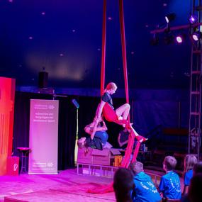 Zwei Akrobatinnen zeigen eine Partnerakrobatiknummer mit roten Tuchbändern in einem Zirkuszelt. Zuschauer sitzen im Hintergrund.