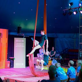 Zwei Artistinnen führen eine Luftakrobatik-Nummer mit roten Tüchern in einem Zirkuszelt auf. Zuschauer beobachten gespannt.