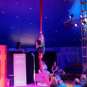 Mädchen schwingt an rotem Tuch in einem Zirkuszelt, während Zuschauer davor gebannt zuschauen.