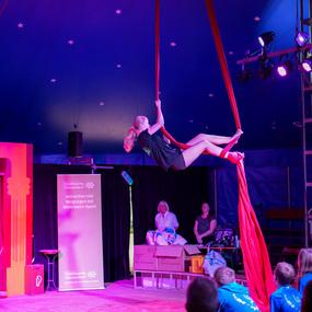 Akrobatin schwingt an roten Tüchern in einem Zirkuszelt vor Publikum, während bunten Lichtern auf die Darbietung scheinen.