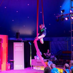 Akrobatin schwingt an roten Tüchern in einem Zelt, Publikum im Hintergrund applaudiert. Farbig beleuchtet.