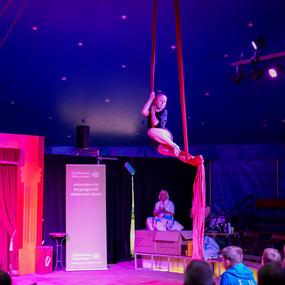 Akrobatin schwingt an roten Tüchern in einem Zelt, während das Publikum zuschaut. Bunte Beleuchtung und Bühne sichtbar.