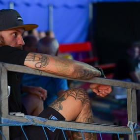 Junger Mann mit Tattoos sitzt entspannt an einer Metallbarriere in einer Zirkus- oder Veranstaltungshalle.