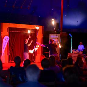 Zwei Künstler führen in einem Zirkuszelt eine Feuerjonglage vor, während das Publikum zuschaut.