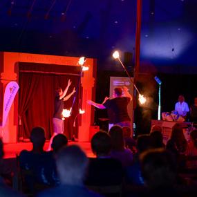 Zwei Artisten führen im Zirkuszelt mit brennenden Fackeln eine Feuershow vor einem Publikum auf.