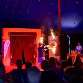 Zwei Artisten führen eine Feuershow in einem Zelt vor Publikum, das gespannt zusieht.