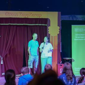 Zwei Personen stehen auf einer Bühne im Circus ZappZarap und sprechen ins Mikrofon, Publikum schaut zu.