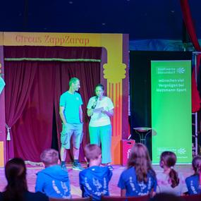 Zwei Personen stehen vor einem Vorhang auf der Bühne eines Zirkus, während Kinder im Hintergrund zuhören.