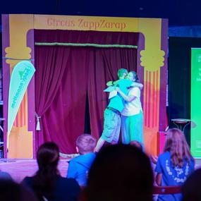 Zwei Personen umarmen sich auf der Bühne des Zirkus ZappZarap vor einem roten Vorhang, während Kinder im Publikum zuschauen.
