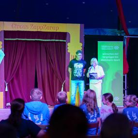 Zwei Personen stehen auf einer Bühne im Circus ZappZarap und sprechen zu einem Publikum aus Kindern.