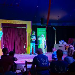 Zwei Personen auf einer Bühne im Zirkuszelt während einer Veranstaltung, Zuschauer sitzen auf Holzstühlen.