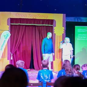Zwei Männer stehen auf einer Bühne im Zirkuszelt vor einem roten Vorhang, während Zuschauer in blauen T-Shirts zuschauen.
