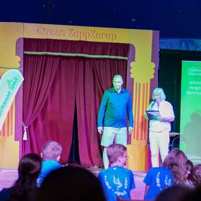Zwei Personen stehen auf einer Bühne im Circus ZappZarap, während das Publikum sie beobachtet. Colorierte Hintergründe sind sichtbar.