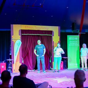 Zwei Personen auf der Bühne des Circus ZappZarap, während eine dritte im Publikum sitzt. Farbige Lichter und Dekorationen sichtbar.