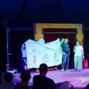 Personen halten ein großes, bemaltes Banner auf einer Zirkusbühne, während das Publikum im Hintergrund zuschaut.