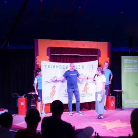 Auf einer Zirkusbühne präsentieren vier Personen ein großes Plakat mit einer bunten Zeichnung und dem Schriftzug „TRIANDER“.