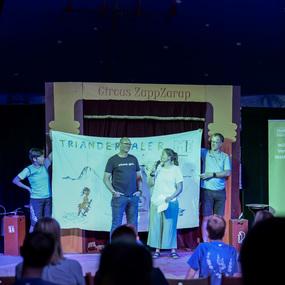 Vier Personen stehen auf der Bühne eines Zirkus mit einem Banner, das eine Zeichnung und den Text "TRIANDER" zeigt.