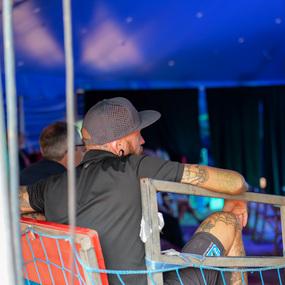 Mann mit Tätowierungen und Cap sitzt in einem Zelt und blickt auf eine Veranstaltung vor ihm.