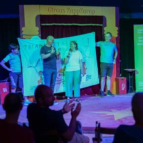 Zwei Personen halten ein Plakat auf einer Bühne im Zirkus, während Zuschauer im Hintergrund applaudieren.