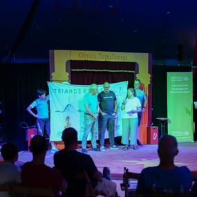 Vier Personen stehen auf einer Zirkusbühne vor einem Banner mit „TRIANDERTIME“, während das Publikum zuschaut.