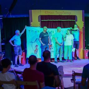 Auf einer Bühne stehen mehrere Personen, die ein großes Banner präsentieren und miteinander sprechen.