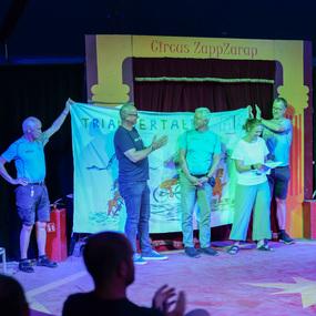 Vier Personen stehen auf einer Zirkusbühne, halten ein Banner und interagieren mit dem Publikum.