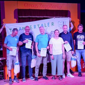 Eine Gruppe von sieben Personen steht auf einer Bühne, hält Urkunden und posiert vor einem Banner im Zirkus.