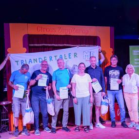 Gruppenszene von Teilnehmern mit Urkunden und Geschenken in einem Zirkus, Banner mit „TRIANDE RTALER“ im Hintergrund.
