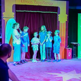 Sechs Kinder stehen auf einer Zirkusbühne, während eine Frau mit Mikrophon spricht, bunte Lichter im Hintergrund.