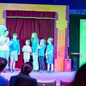 Eine Moderatorin liest vor, während mehrere Kinder auf einer Bühne im Zirkuszelt stehen, umgeben von bunten Kulissen.