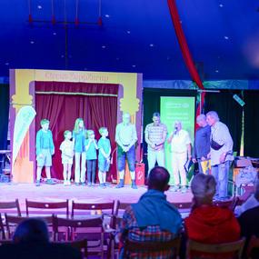 Bühne im Zirkuszelt mit Kindern und Erwachsenen, die sich versammelt haben, während ein Sprecher spricht.