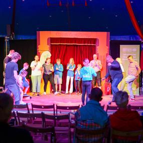 Bunte Zirkusaufführung mit Kindern und Erwachsenen auf der Bühne, umgeben von Publikum in einem festlichen Zelt.