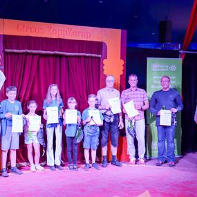 Gruppenfoto von acht Personen, die Urkunden in einem Zirkuszelt halten, mit rotem Vorhang im Hintergrund.