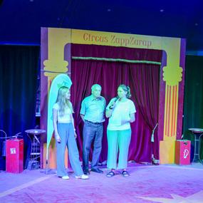 Zwei Frauen und ein Mann stehen auf einer Zirkusbühne und sprechen ins Mikrofon, umgeben von bunten Vorhängen.