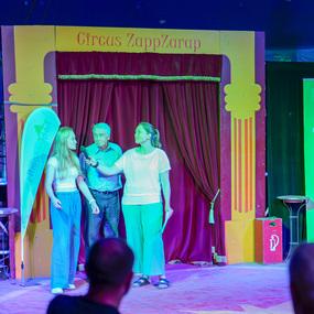 Drei Personen stehen auf einer Bühne im Zirkuszelt, eine spricht ins Mikrofon, farbige Beleuchtung im Hintergrund.