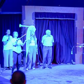 Aufführung im Zirkus mit mehreren Personen, die Jonglierstäbe halten, vor einem Vorhang in blauer Beleuchtung.