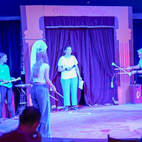 Zwei junge Frauen und ein älterer Mann stehen auf einer Bühne im Zirkus, während sie mit jonglierenden Requisiten arbeiten.