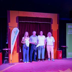 Vier Personen posieren vor einem Bühnenvorhang im Zirkus ZappZarap, halten Urkunden und lächeln.