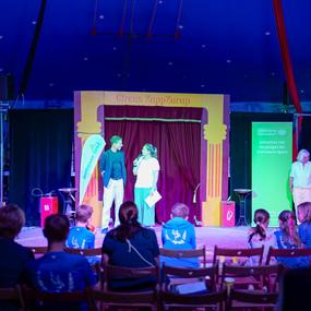 Zwei Personen stehen auf einer Bühne im Zirkuszelt und sprechen miteinander, während das Publikum zuschaut.