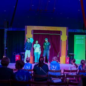 Drei Personen stehen auf einer Bühne im Zirkuszelt, während das Publikum aufmerksam zuschaut.